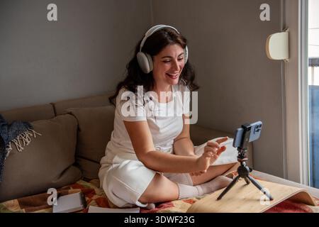 Una donna sorridente si siede su un comodo divano di casa, coinvolgendo il suo pubblico tramite uno smartphone mentre indossa le cuffie. Lei crea contenuti in un relax Foto Stock