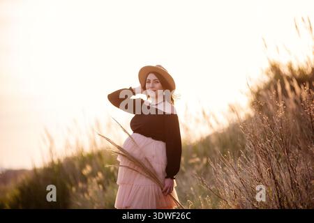Una giovane donna incinta si trova graziosamente in un prato illuminato dal sole, abbracciando la natura con un sorriso dolce. Indossa un cappello elegante e scorre con la calma sur Foto Stock