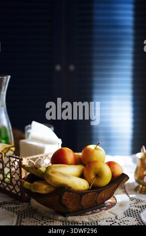 Una varietà di frutta fresca, tra cui banane e mele disposte in una ciotola decorativa di legno su un tavolo domestico, evidenziando il tema di un'alimentazione sana. Foto Stock