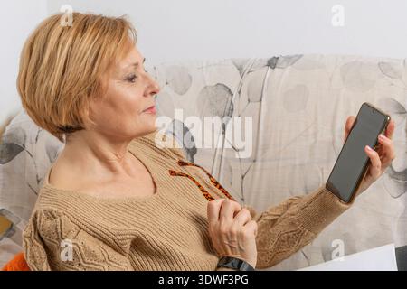 Donna bionda di mezza età che controlla il suo smartphone seduto sul divano di casa Foto Stock