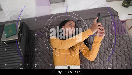 Donna sdraiata in pantaloni leggeri con parte superiore della senape che regge lo smartphone sul letto, con valigia, passaporto e HUD Foto Stock