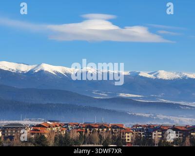 Le montagne innevate si innalzano dietro un tranquillo villaggio di case con tetto rosso, incorniciate da un cielo blu brillante e da morbide nuvole. Il paesaggio invernale evoca calma, trave Foto Stock