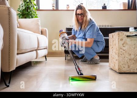 Una donna con un abbigliamento confortevole pulisce il pavimento del suo moderno soggiorno utilizzando un aspirapolvere senza filo. La luce naturale scorre attraverso la finestra, evidenziando Foto Stock