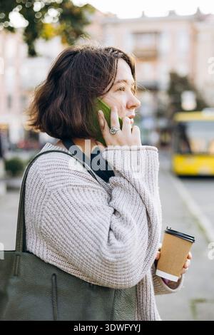 Una donna che indossa un comodo maglione parla al telefono cellulare, tenendo una tazza di caffè all'aperto. Si trova vicino a una strada con un veicolo giallo nella ba sfocata Foto Stock