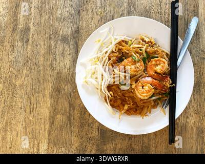 Cospargere Thai con gamberi, germogli di soia e lime presentati in una ciotola bianca, accompagnati da bacchette, posti su un rustico tavolo di legno Foto Stock