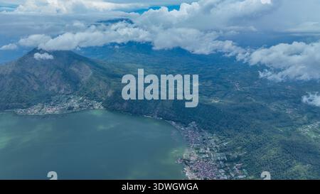 Il lago color smeraldo della Caldera di Batur si snoda con grazia lungo scogliere spettacolari, abbracciando borghi lacustri tra foreste verdeggianti e vette lontane in un'area aerea serena Foto Stock