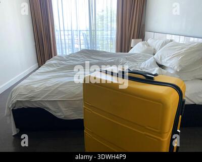 La valigia gialla si erge accanto a un letto non fatto in una camera d'hotel, caratterizzata da una grande finestra con tende a vista e un'atmosfera luminosa e ariosa Foto Stock