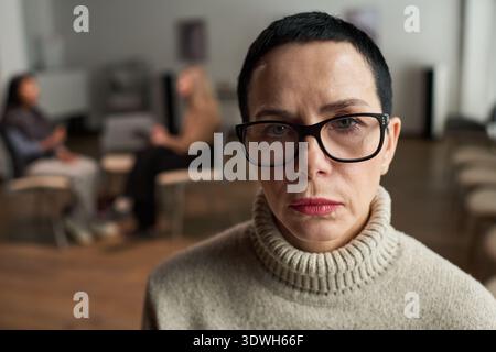Ritratto di una donna caucasica di mezza età triste con capelli corti e occhiali che guardano nella fotocamera, seduta in primo piano mentre due donne conversano in uno sfondo sfocato Foto Stock