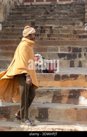 Venditore di Chai avvolto in abiti caldi che camminano su gradini di pietra a Varanasi, in India, vicino al suo set di tè. Foto Stock