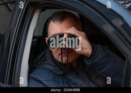 Un uomo siede in un'auto guardando attraverso un binocolo fuori dal finestrino del veicolo, sorvegliando sotto copertura e monitorando le strade in un ambiente urbano Foto Stock