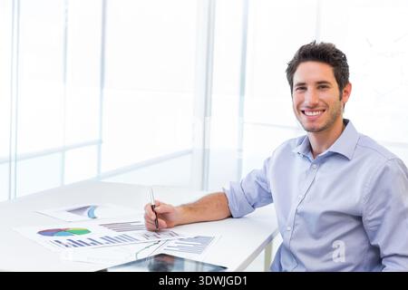 Uomo seduto alla scrivania dell'ufficio con una camicia blu che tiene in mano una penna che esamina i referti stampati, il tablet e lo spazio per le copie. Minimalista, moderno, luminoso, pulito, dati, grafici, Foto Stock