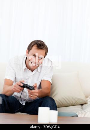 Uomo seduto a casa sul divano che indossa una camicia bianca e jeans scuri con controller cablato. Gaming, salotto, candele, neutro, accogliente, ambiente, informale Foto Stock