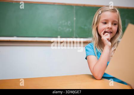 Bambina seduto alla scrivania della classe con carta abbronzante e dietro la tavola blu, spazio per fotocopie. Lavagna, studente, focus, istruzione, studio, biondo Foto Stock