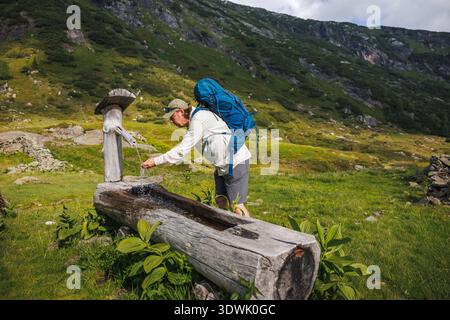 Acqua rinfrescante proveniente dalla sorgente di montagna. Escursionista assetato che beve acqua dalla fontana tradizionale durante il viaggio di trekking nelle Alpi. Fonte d'acqua naturale Foto Stock