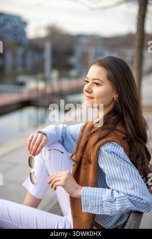 Giovane donna elegante in stile vecchio denaro seduta sulla panchina vicino al molo di Praga con gli occhiali da sole all'inizio della primavera. Lusso discreto e senza tempo Foto Stock