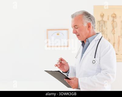 Medico senior che esamina gli appunti in clinica indossando cappotto e stetoscopio, tenendo la penna, spazio per le copie. Medico, ufficio, certificato, anatomico, scheletrico, Foto Stock