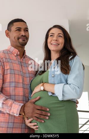 Diverse coppie che si aspettano di mettere le mani su un batuffolo e stare in piedi all'interno di casa vicino alla ringhiera delle scale. Gravidanza, famiglia, maternità, aspettativa, casa, relazione, calore || modello rilasciato Foto Stock