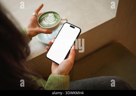 Vista dall'alto di una giovane donna seduta a un tavolo in un bar che beve un latte matcha caldo mentre utilizza un telefono cellulare con uno schermo vuoto Foto Stock