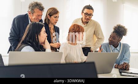 Un gruppo eterogeneo di dirigenti e leader aziendali multigenerazionali si riuniscono intorno ai notebook in un ufficio luminoso per discutere di pianificazione strategica e realizzazione Foto Stock