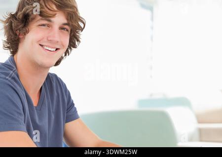 Uomo adulto seduto a sinistra in camera, sorridente, con scollo a V navy, seduta di tè dietro, spazio fotocopie. Soleggiato, minimalista, moderno, lounge, allegro, candi Foto Stock