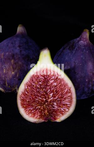 Tre fichi viola maturi con uno tagliato a metà per rivelare polpa e semi rossi vivaci su uno sfondo nero drammatico Foto Stock