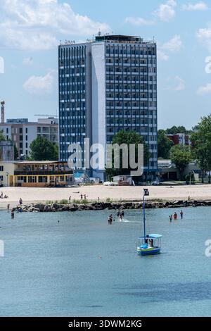 La gente cammina lungo la spiaggia e nuota in acqua in una giornata di sole. Una piccola barca è sull'acqua vicino a un alto edificio e ad altre strutture Foto Stock