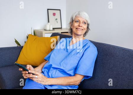 Lavoratore sanitario senior che si rilassa sul divano utilizzando il telefono cellulare dopo la giornata lavorativa Foto Stock