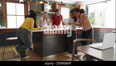 Un gruppo di sei adulti che esamina un documento sulla moderna isola della cucina, tenendo tazze e laptop Foto Stock