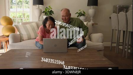 Donna pendente con top rosa e uomo con camicia verde che utilizza un laptop su un tavolo di legno a casa, mostrando dati numerici Foto Stock