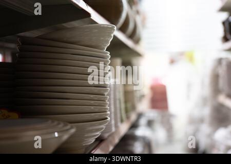 Piastre in ceramica bianca impilate sul ripiano nell'area di stoccaggio della cucina, sfondo sfocato con illuminazione soffusa Foto Stock