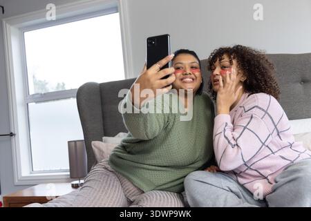Diverse amiche che scattano selfie con lo smartphone a letto, indossando toppe sotto gli occhi e pigiami Foto Stock