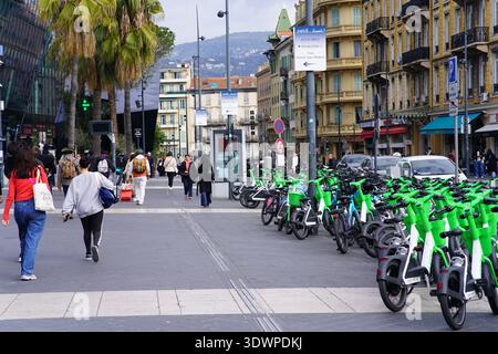 Pedoni che camminano lungo la strada fiancheggiata da palme nel centro di Nizza con scooter elettrici verdi Lime parcheggiati, scena urbana della Costa Azzurra, Costa Azzurra Francia. Foto Stock