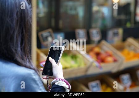 Una giovane donna in giacca blu cattura una vivace bancarella di frutta traboccante di banane, mele, arance e pesche sul suo smartphone in un mercato di Nizza. Foto Stock