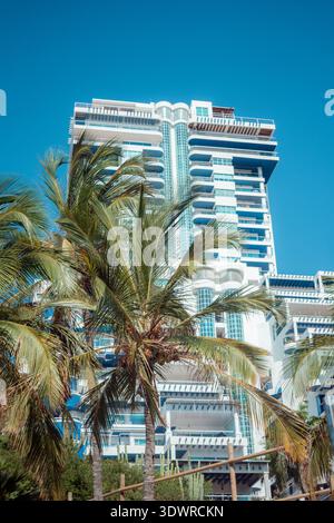 Moderno ed alto edificio alberghiero in una destinazione tropicale con elementi architettonici bianchi e blu e numerosi balconi, incorniciati da vibranti giardini verdi Foto Stock
