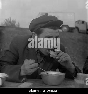 Lo storico ritratto in bianco e nero degli anni '1970 cattura un autista o un lavoratore agricolo durante una pausa pranzo presso l'impresa statale Donrybkombinat nel distretto di Slavyansk, RSS Ucraina, URSS. Indossato con un classico cappello piatto e una giacca, viene mostrato mentre muta un pezzo di pane mentre mangia un pasto semplice. Sullo sfondo, il suo furgone bianco è parzialmente visibile sul fianco della collina. Questa autentica fotografia d'archivio mette in evidenza la routine quotidiana della classe operaia sovietica e l'atmosfera ottimistica nel tranquillo Donbass prima della guerra. Foto Stock