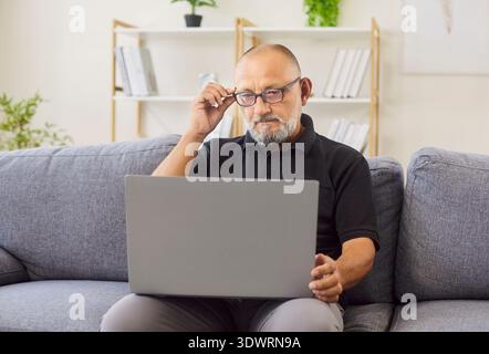 Un uomo maturo regola gli occhiali mentre lavora su un computer portatile all'interno del salotto di casa Foto Stock