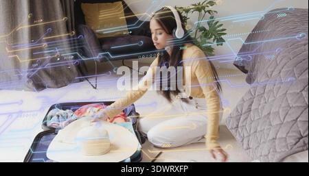 Donna inginocchiata che indossa una valigia rigida in camera da letto, indossa la mostarda, cuffie, mascherina digitale Foto Stock