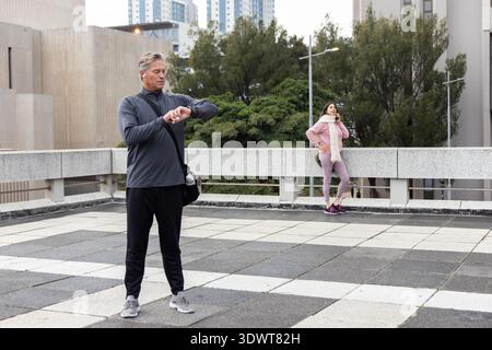 Uomo anziano in abbigliamento sportivo con borsa a tracolla che controlla l'orologio sulla terrazza sul tetto, donna che parla al telefono Foto Stock