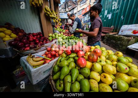 Vari tipi di frutta fresca sono esposti in una vivace banchina del mercato nel Bazaar inferiore a Shimla, con persone ed edifici visibili in lontananza. Foto Stock