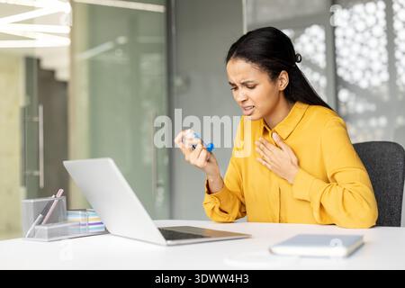 Giovane donna che soffre di asma alla sua scrivania, stringendosi il petto e usando un inalatore mentre lavora su un notebook in un ufficio moderno, in difficoltà e senza fiato Foto Stock