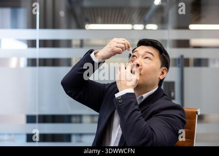 Uomo asiatico in abbigliamento da lavoro alla scrivania dell'ufficio che applica gocce agli occhi irritati, gestisce l'irritazione quotidiana degli occhi e pratica l'autoassistenza mentre lavora Foto Stock