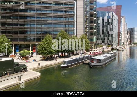 Le persone si rilassano vicino al canale di Paddington Basin, Londra, con barche strette ormeggiate vicino all'architettura moderna. Foto Stock
