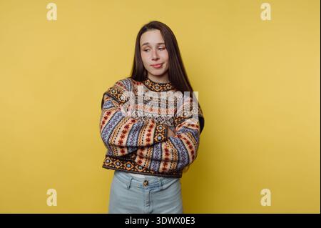 Una giovane donna sta in piedi con le braccia incrociate e indossa un comodo maglione a motivi geometrici. L'allegro sfondo giallo mette in risalto la sua atmosfera divertente e rilassata Foto Stock