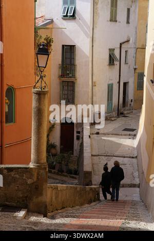 Una coppia di anziani cammina lungo i vicoli stretti e colorati con ripidi scalini acciottolati nel vecchio villaggio. Mentone, Francia Foto Stock