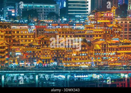Chongqing, Cina - 8 dicembre 2025: Edifici illuminati in stile tradizionale della grotta di Hongya si innalzano su livelli sopra la trafficata strada lungo il fiume. Storico cinese Foto Stock