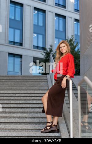 Una donna bionda che indossa una camicia rossa e una gonna marrone posa su una rampa di gradini in cemento in un ambiente urbano. Un grande e moderno edificio e' visibile in Foto Stock