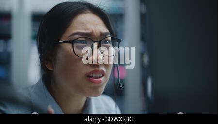 Il trader asiatico riceve la chiamata tramite auricolare mentre monitora i dati finanziari presso la moderna Borsa valori. Donna ascolta le cattive notizie sulle prestazioni del mercato, l'espressione sposta preoccupazione e sofferenza. Foto Stock