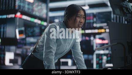 Emotional Asian Woman Broker riceve notizie devastanti sulla cuffia sull'improvviso crollo del mercato azionario. La donna arrabbiata si alza nelle carte Falling rosse della scansione panica. Stress professionale e crisi finanziaria. Foto Stock