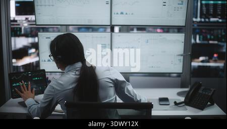 Commerciante di cuffie che lavora in una workstation sofisticata. La donna interagisce con il tablet digitale monitorando quattro grandi schermi pieni di complessi grafici del mercato azionario, analisi in tempo reale. Vista posteriore. Foto Stock