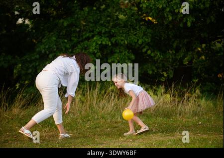 La ragazza e sua madre giocheranno felicemente con la palla sul campo, correndo felicemente con essa. Madre e figlia giocano insieme fuori durante le vacanze. Concetto: Infanzia, sogni, giochi divertenti Foto Stock